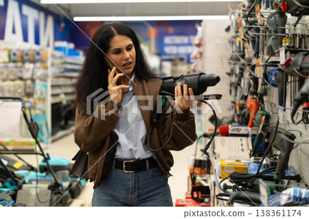 Woman in hardware store holding power drill while talking on smartphone 138361174