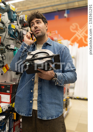 Man talking on phone while holding a circular saw in a tools shop Man talking on phone while holding a circular saw in a tools shop 138361291
