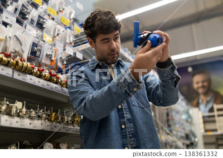 Young man shopping for plumbing components, examining a blue valve in a diy store Young man shopping for plumbing components, examining a blue valve in a diy store 138361332