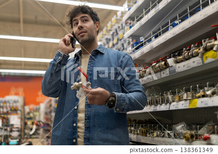 Man talking on smartphone holding a valve, shopping for plumbing supplies in a hardware store Man talking on smartphone holding a valve, shopping for plumbing supplies in a hardware store 138361349