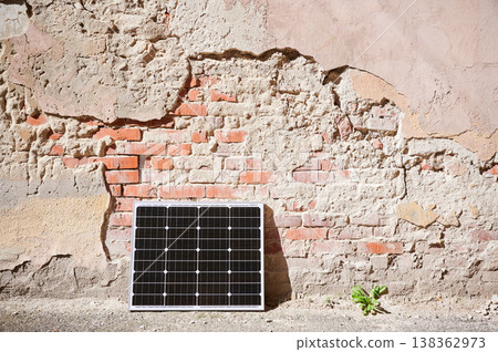 Photovoltaic solar panel at weathered, textured wall with peeling paint. Juxtaposition of modern renewable energy technology against rustic, decaying backdrop highlights contrast between old and new. Photovoltaic solar panel at weathered, textured wall with peeling paint. Juxtaposition of modern renewable energy technology against rustic, decaying backdrop highlights contrast between old and new. 138362973