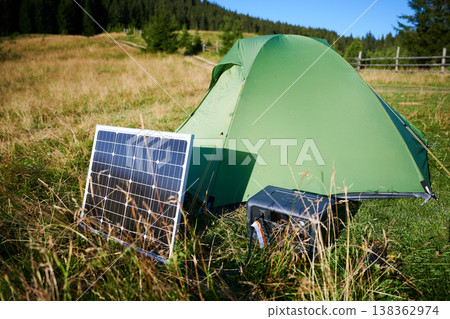 Tourist tent with solar panel and portable power station nearby in grassy field. Scene surrounded by rolling hills, showcasing eco-friendly camping setup. Tourist tent with solar panel and portable power station nearby in grassy field. Scene surrounded by rolling hills, showcasing eco-friendly camping setup. 138362974