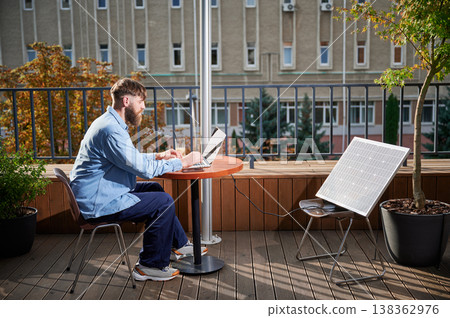 Man works on laptop at outdoor round table, with glass of iced coffee. Solar panel nearby, emphasizing sustainable, modern eco-friendly workspace that combines technology and sustainability. Man works on laptop at outdoor round table, with glass of iced coffee. Solar panel nearby, emphasizing sustainable, modern eco-friendly workspace that combines technology and sustainability. 138362976
