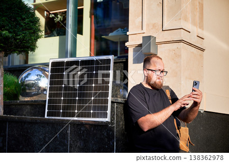 Bearded man using smartphone next to photovoltaic solar panel. Integration of sustainable renewable energy into everyday life, demonstrating practical use of solar power for charging devices. Bearded man using smartphone next to photovoltaic solar panel. Integration of sustainable renewable energy into everyday life, demonstrating practical use of solar power for charging devices. 138362978