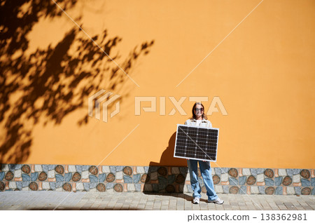 Woman holding solar panel for charging, stands against vibrant orange wall. Smiling girl wears sunglasses, white top, blue jeans. Artistic shadows cast by nearby trees branches on wall. Woman holding solar panel for charging, stands against vibrant orange wall. Smiling girl wears sunglasses, white top, blue jeans. Artistic shadows cast by nearby trees branches on wall. 138362981