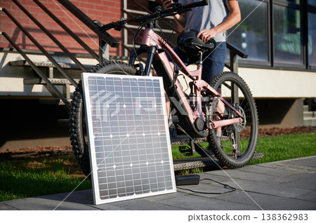 Pink electric mountain bike connected to charging device with solar panel. Man cyclist stands on staircase beside brick building, using smartphone. Focus on solar panel. Pink electric mountain bike connected to charging device with solar panel. Man cyclist stands on staircase beside brick building, using smartphone. Focus on solar panel. 138362983