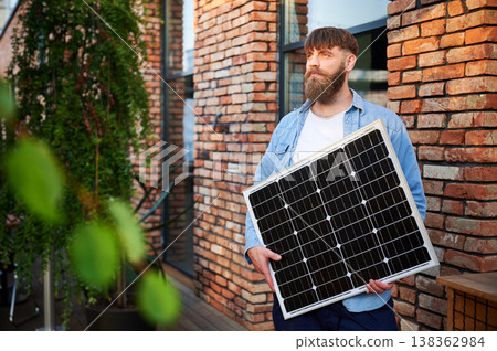 Portrait of bearded man holds solar panel, standing in front of brick building and lush greenery. Guy in shirt and t-shirt exudes calm and eco-conscious vibe, blending nature with urban elements. Portrait of bearded man holds solar panel, standing in front of brick building and lush greenery. Guy in shirt and t-shirt exudes calm and eco-conscious vibe, blending nature with urban elements. 138362984