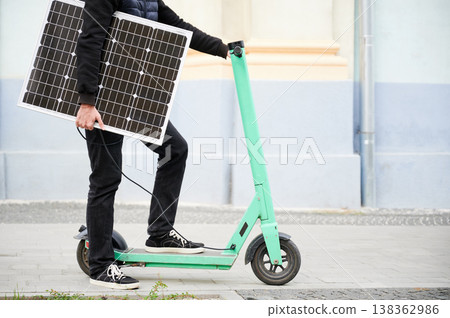 Cropped view of man rides electric scooter while carrying solar panel. Integration of solar power as sustainable energy source for charging electric scooters, promoting eco-friendly transportation. 138362986