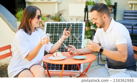 Man and woman charging smartphone from solar panel, relaxing in cafe. Cute couple using portable solar charger for devices. Charming couple sitting on terrace on background of solar battery. Man and woman charging smartphone from solar panel, relaxing in cafe. Cute couple using portable solar charger for devices. Charming couple sitting on terrace on background of solar battery. 138362988