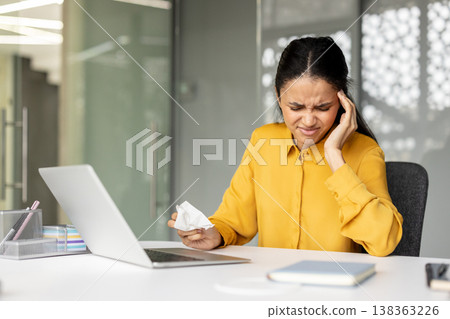 Young business woman feeling intense headache or migraine, holding her temple and a tissue, sitting at a laptop in a modern office setup, experiencing work-related stress, burnout, or illness 138363226