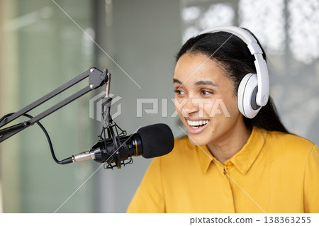 Young woman wearing headphones and speaking into a microphone, hosting a live podcast from her home studio, smiling while creating engaging audio content and streaming online Young woman wearing headphones and speaking into a microphone, hosting a live podcast from her home studio, smiling while creating engaging audio content and streaming online 138363255