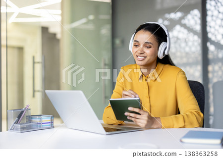 Young woman wearing headphones and a yellow shirt, smiling and writing notes in a small book while attending an online meeting or webinar using a laptop at a modern office desk 138363258