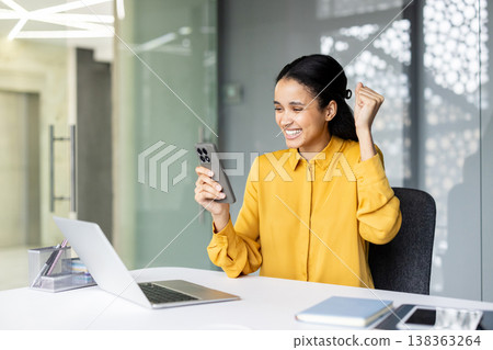 Happy young woman sitting at a desk in an office, celebrating an achievement or good news while looking at her smartphone and raising a fist in triumph Happy young woman sitting at a desk in an office, celebrating an achievement or good news while looking at her smartphone and raising a fist in triumph 138363264