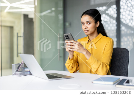 Young professional woman using a smartphone in a modern, bright office with a laptop on the desk, focusing on digital communication and multitasking during her workday 138363268
