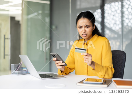 Stressed woman experiencing a credit card payment problem on a mobile phone, trying to make an online purchase while sitting at an office desk with a laptop 138363270