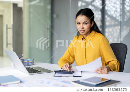 Young woman accountant wearing a yellow shirt and calculating financial data using a calculator and papers, while working at her desk in a contemporary office setting 138363273