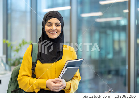 Muslim student woman wearing a hijab and backpack, confidently smiling while holding notebooks, representing education, success, diversity, and modern learning in a blurred university setting Muslim student woman wearing a hijab and backpack, confidently smiling while holding notebooks, representing education, success, diversity, and modern learning in a blurred university setting 138363290