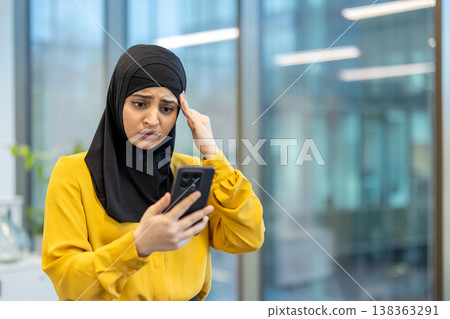 Muslim businesswoman in a hijab and yellow blouse looks worried and stressed while reading bad news on her smartphone in a modern office, facing a workplace crisis 138363291