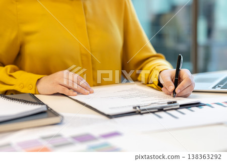 Business person in yellow shirt sitting at office desk, holding a pen and accurately signing a paper document on a clipboard during important contract work Business person in yellow shirt sitting at office desk, holding a pen and accurately signing a paper document on a clipboard during important contract work 138363292