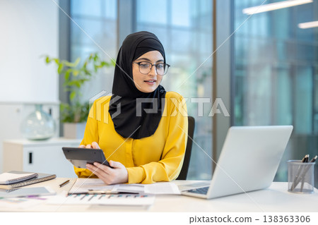Muslim woman wearing a hijab and glasses, working on financial calculations with a digital calculator and a laptop at a modern office desk, demonstrating professionalism and expertise 138363306