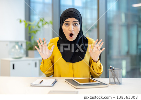 Muslim business woman wearing a black hijab and yellow shirt sitting at an office desk, reacting with wide eyes and an open mouth, showing shock and amazement 138363308