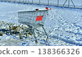 A silver metal shopping cart with red rims parked in the snow near a sidewalk. 138366326