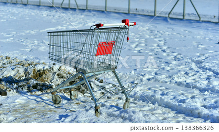 A silver metal shopping cart with red rims parked in the snow near a sidewalk. 138366326