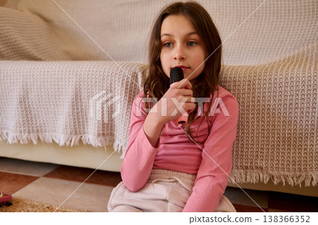 Young Girl Singing Into Microphone At Home On Sofa In Casual Pink Outfit Young Girl Singing Into Microphone At Home On Sofa In Casual Pink Outfit 138366352