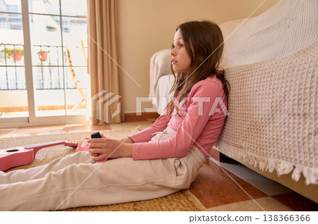 Young Girl Sitting on Living Room Floor With Pink Guitar Looking Thoughtful 138366366