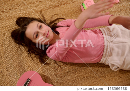 Young Girl Lying on Woven Rug Reaching Up Beside Pink Ukulele and Headphones Young Girl Lying on Woven Rug Reaching Up Beside Pink Ukulele and Headphones 138366388
