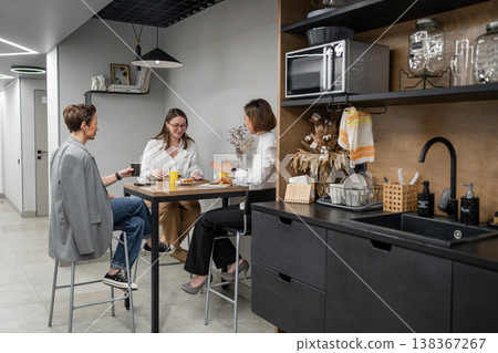 Women talking and dining at kitchen island counter in open office lunch area with tableware. Dining, kitchen, talking, teamwork, office, women. Women talking and dining at kitchen island counter in open office lunch area with tableware. Dining, kitchen, talking, teamwork, office, women. 138367267