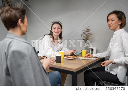 Women eating, smiling, and talking around table in company kitchen during lunch break. Eating, lunch, kitchen, teamwork, smiles, office. Women eating, smiling, and talking around table in company kitchen during lunch break. Eating, lunch, kitchen, teamwork, smiles, office. 138367270