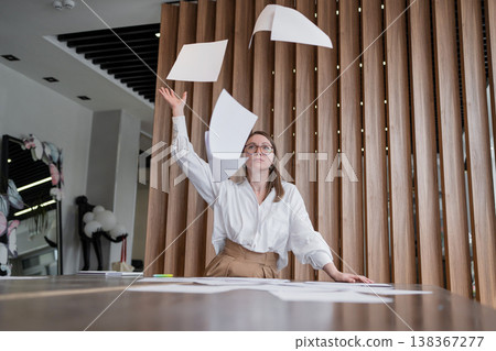 Woman joyfully tossing paper sheets in the air at desk in modern office with wooden wall. Tossing, paper, desk, air, joy, woman, office. Woman joyfully tossing paper sheets in the air at desk in modern office with wooden wall. Tossing, paper, desk, air, joy, woman, office. 138367277