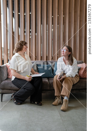 Coworkers sitting on couch talking and exchanging ideas beside wood panel wall at office. Talking, exchanging, ideas, couch, coworkers, office. Coworkers sitting on couch talking and exchanging ideas beside wood panel wall at office. Talking, exchanging, ideas, couch, coworkers, office. 138367279