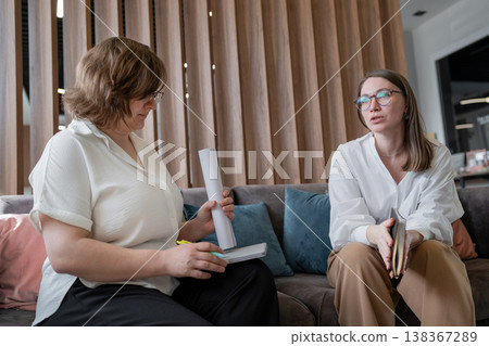 Female team members consulting and smiling with papers on sofa in stylish office lounge. Consulting, smiling, sofa, team, office, women. Female team members consulting and smiling with papers on sofa in stylish office lounge. Consulting, smiling, sofa, team, office, women. 138367289