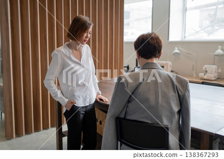 Businesswoman approaching and greeting colleague at desk in sunny modern workspace. Greeting, desk, sunny, workspace, businesswoman, colleague. Businesswoman approaching and greeting colleague at desk in sunny modern workspace. Greeting, desk, sunny, workspace, businesswoman, colleague. 138367293