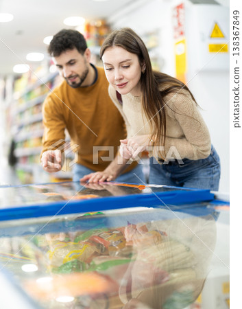 Positive young couple choosing cooled food product standing at fridge in grocery store Positive young couple choosing cooled food product standing at fridge in grocery store 138367849