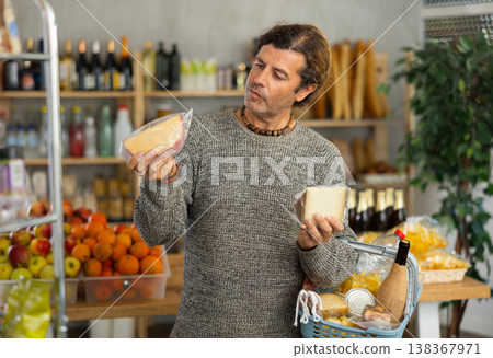 Attentive man with shopping basket trying to make choice holding two sorts of cheese in grocery store Attentive man with shopping basket trying to make choice holding two sorts of cheese in grocery store 138367971