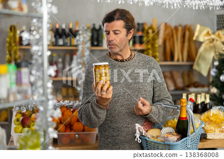 Positive male shopper with shopping basket choosing olives for New Year's holiday at grocery store Positive male shopper with shopping basket choosing olives for New Year's holiday at grocery store 138368008
