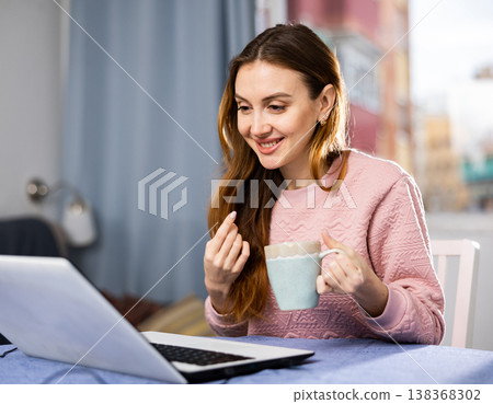 Portrait of young woman sitting at table in living room looking laptop screen Portrait of young woman sitting at table in living room looking laptop screen 138368302