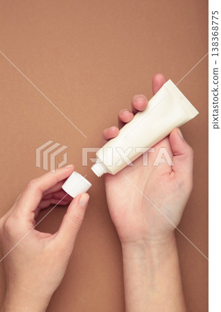 Beige cream tube in woman hands on brown background. Skin care concept. 138368775