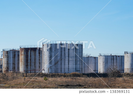 Row of large industrial storage tanks with rust and weathering in open field under blue sky Row of large industrial storage tanks with rust and weathering in open field under blue sky 138369131