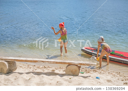 Girls in swimsuits are having fun on the shore of the lake, splashing water from a bucket. Summer vacation vacation. Girls in swimsuits are having fun on the shore of the lake, splashing water from a bucket. Summer vacation vacation. 138369204