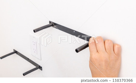 Black metal shelf bracket held against white wall showing L-shape and screw holes. Home improvement, hardware presentation, DIY shelf bracket, wall mounting prep. Black metal shelf bracket held against white wall showing L-shape and screw holes. Home improvement, hardware presentation, DIY shelf bracket, wall mounting prep. 138370366