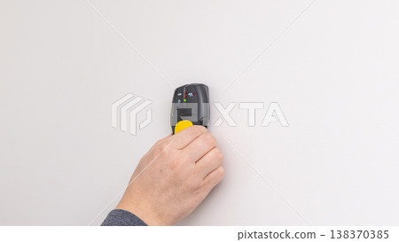 Mans hand pressing yellow and black stud finder onto white wall close to power outlet. Home improvement, detecting studs, DIY process, tool application. 138370385