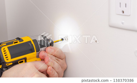 Mans hands using yellow cordless drill with light on, illuminating wall area near outlet during work. Home improvement, drilling with flashlight, DIY task, illuminated workspace. 138370386