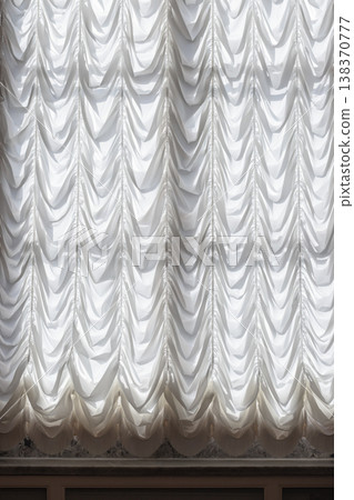 Elegant white drapery with gathered swags cascades over a backlit window Elegant white drapery with gathered swags cascades over a backlit window 138370777