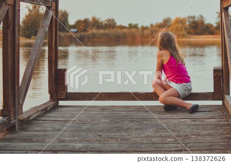 A cute young girl sits on a wooden pier at the end of a lake. The photo was taken during the golden hour, at sunset. It represents childhood, summer vacations, looking toward the future, and carefree A cute young girl sits on a wooden pier at the end of a lake. The photo was taken during the golden hour, at sunset. It represents childhood, summer vacations, looking toward the future, and carefree 138372626