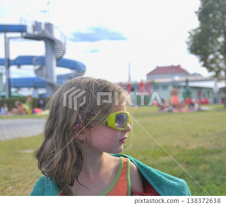 A cute little girl wearing sunglasses at a swimming pool. A smiling little girl wrapped in a towel at a swimming pool with a water slide in the background. The concept of summer, summer vacation, A cute little girl wearing sunglasses at a swimming pool. A smiling little girl wrapped in a towel at a swimming pool with a water slide in the background. The concept of summer, summer vacation, 138372638