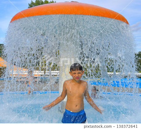 A boy under a waterfall at a public swimming pool. A little boy under a sprinkler at a swimming pool. Copy space. The concept of summer, summer holidays, free time, and relaxation. A boy under a waterfall at a public swimming pool. A little boy under a sprinkler at a swimming pool. Copy space. The concept of summer, summer holidays, free time, and relaxation. 138372641